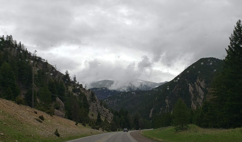 Paved two-lane road winds through a pine valley in Yellowstone National Park, with snow-capped peaks and raindrops on lens.