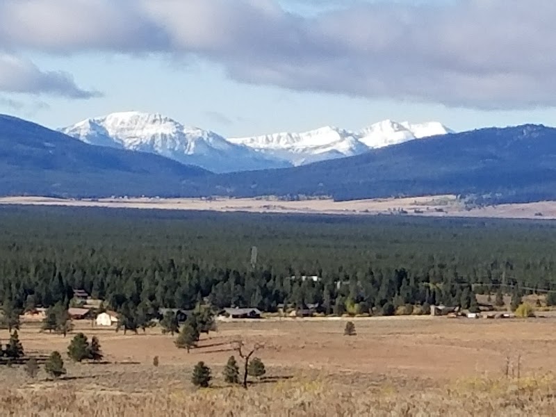 Snow-capped peaks behind a dense pine forest and open meadow with scattered cabins along a Yellowstone National Park trail.