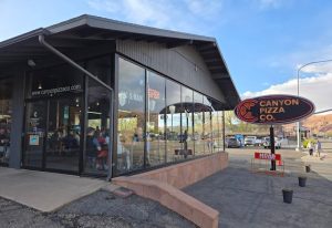 Glass-front restaurant with large windows and outdoor seating, visible parking lot, near Arches National Park.