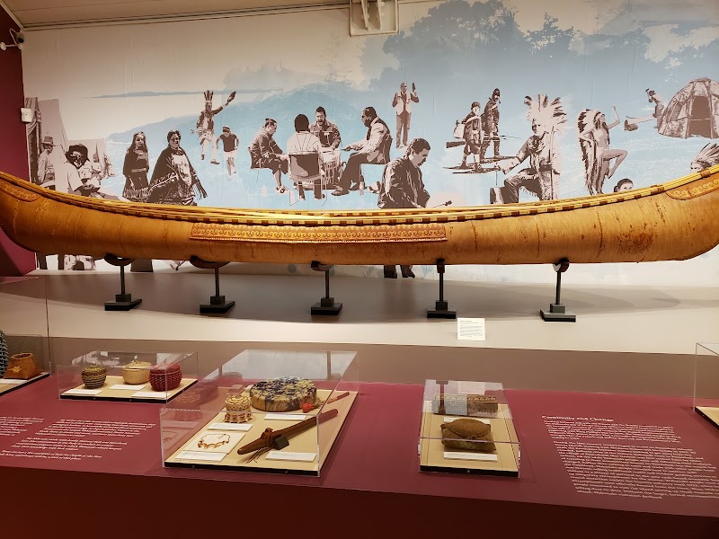 Long wooden canoe on display with a blue mural of people and glass-case artifacts at Acadia National Park.