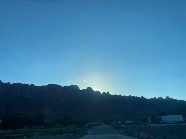 Blue dawn sky over a jagged desert ridge in Arches National Park, with a road and scattered buildings in the foreground.