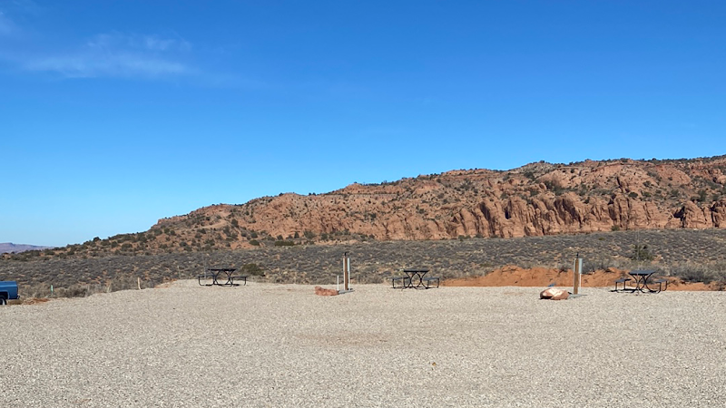 Desert campground at Arches National Park with a gravel lot, a few picnic tables, and red rock cliffs under a clear blue sky.