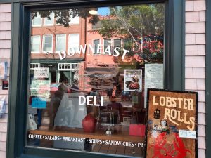 Storefront window of a deli with a lobster rolls signboard, reflections of brick buildings and trees in Acadia National Park.
