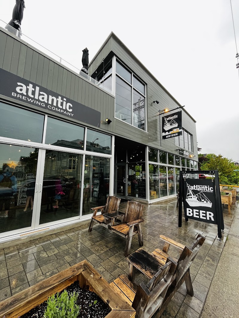 Outside a modern two-story brewery storefront with large glass windows, rustic wooden chairs, and a beer sign in Acadia National Park.