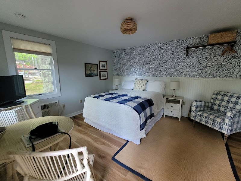 Cozy motel bedroom in Acadia National Park with a white bed and blue plaid throw, patterned wallpaper, two nightstands, wicker chairs, and a TV by the window.