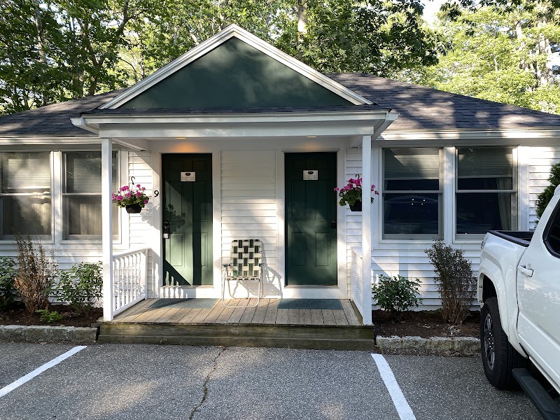 Two green doors on a white motel cottage with a small porch, hanging planters, a metal chair, and a white pickup in Acadia National Park.