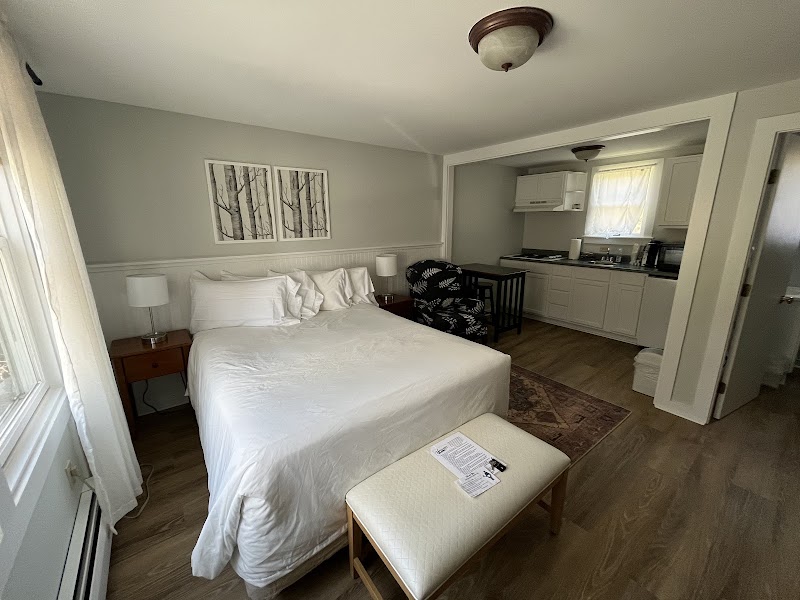 Cozy room with a full bed, white linens, two nightstands, and a bench at the foot; kitchenette visible nearby, Acadia National Park.