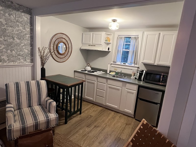 A compact kitchenette with white cabinets, stove, microwave, and mini fridge, plus a bar-height table and striped armchair in Acadia National Park lodging.