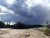 Sour Lake overlooks a barren shore with a pine forest in the distance and dark storm clouds gathering over Yellowstone National Park.