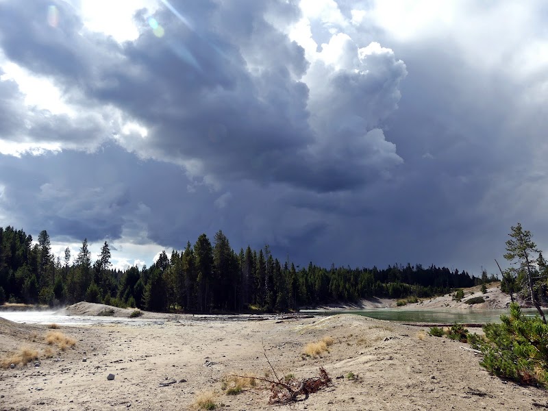 Dark storm clouds over Sour Lake's sandy shore in Yellowstone National Park, with forested hills beyond.