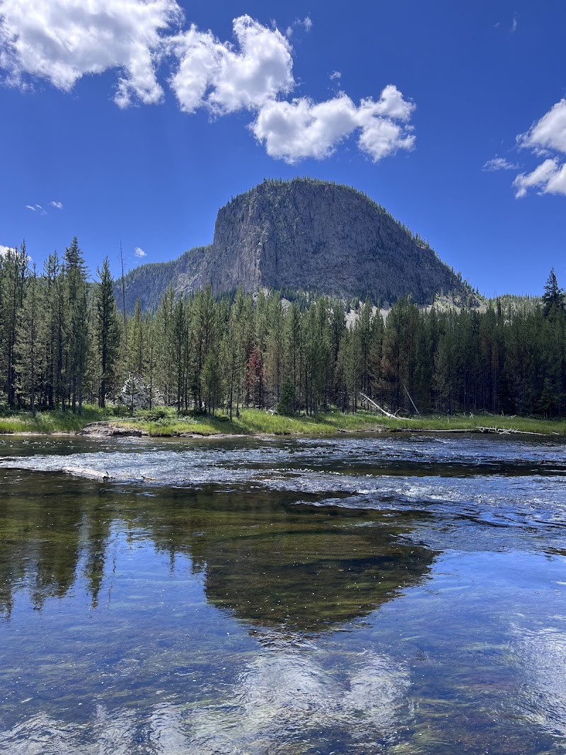 Broad river in foreground reflects a pine forest and a rounded granite peak under blue sky in Yellowstone National Park.