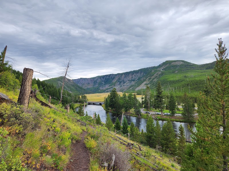 Trail along a grassy hillside in Yellowstone National Park, wooden fence posts, river below, and distant green mountains under a cloudy sky.