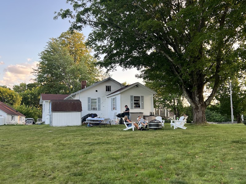 White cottage with gray trim and a large tree in Acadia National Park, with people relaxing on lawn chairs and a grill.