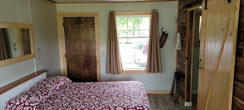Cozy motel room with a red floral comforter on a queen bed, wooden door, window with beige curtains, and a wooden wardrobe in Acadia National Park.