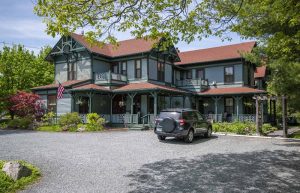 Victorian-style inn with teal siding and a red roof, wraparound porch, flag, and a parked SUV in Acadia National Park.