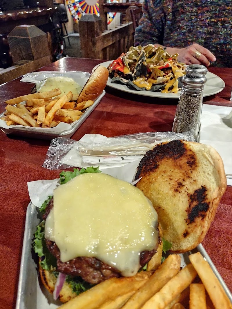 In Yellowstone National Park, a wooden table shows a cheeseburger with melting cheese, lettuce and onion, with fries, and a second plate of loaded nachos nearby.