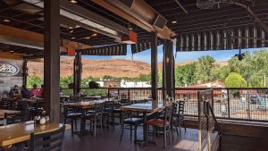 Covered outdoor dining deck at Arches National Park with wooden tables and metal chairs, railing, and red rock hills beyond.
