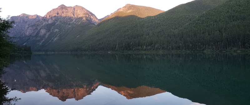 Harrison Lake in Glacier National Park reflects the surrounding peaks and evergreen forest at sunset.