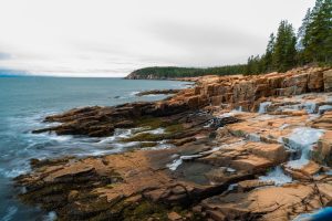 Rugged orange-brown rocks line the Atlantic coast at Thunder Hole in Acadia National Park, with forested cliffs and surf.