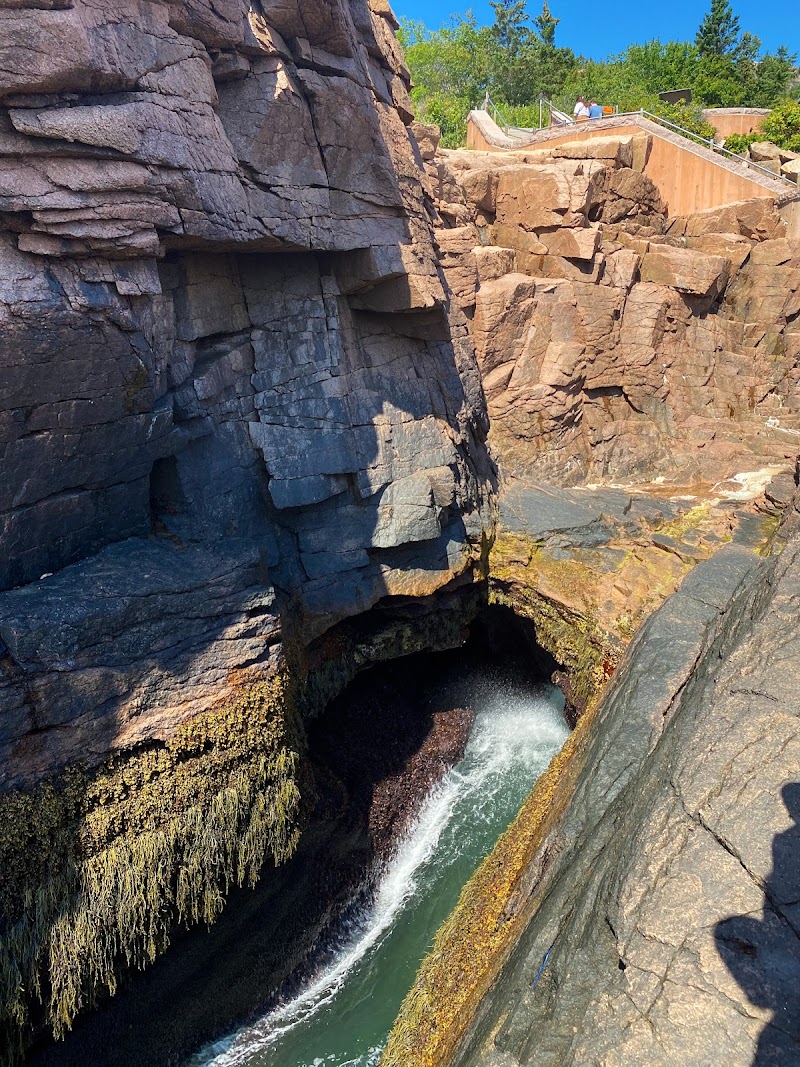 Rugged granite cliffs at Thunder Hole, Acadia National Park, with waves rushing into a dark inlet below a distant boardwalk and green-hued rock faces.