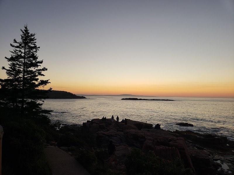 Sunset over a rocky shoreline in Acadia National Park with silhouetted hikers on the rocks and a calm sea.