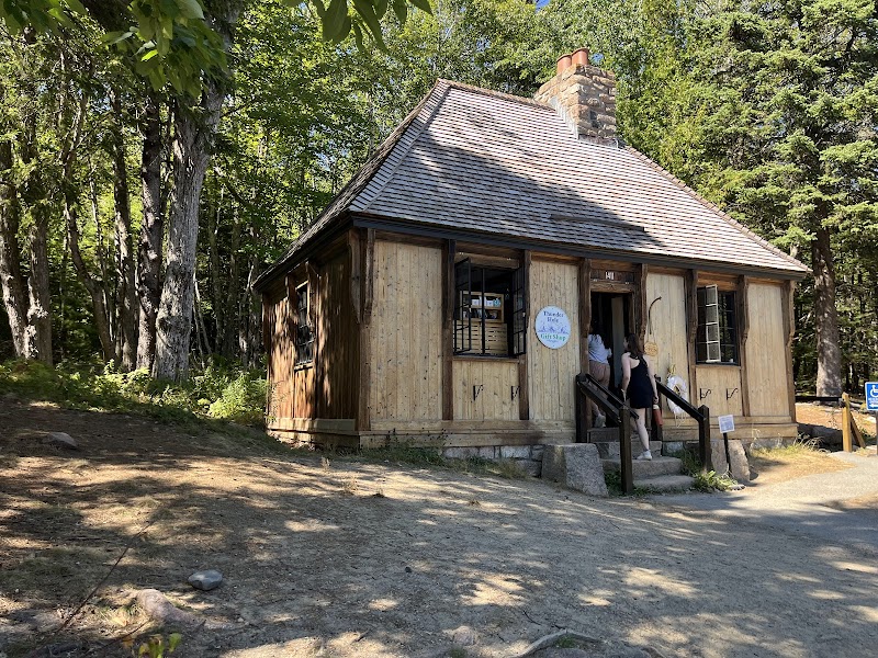 Small wooden cabin with a shingled roof beside tall trees, with two people on the front steps in Acadia National Park.