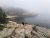 Fog shrouds a rocky shoreline in Acadia National Park as a person walks along pink granite ledges toward the misty sea.
