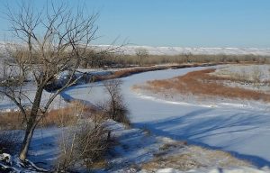 Snowy badlands river bend with bare trees, brown grasses, and distant white ridges in Badlands National Park.