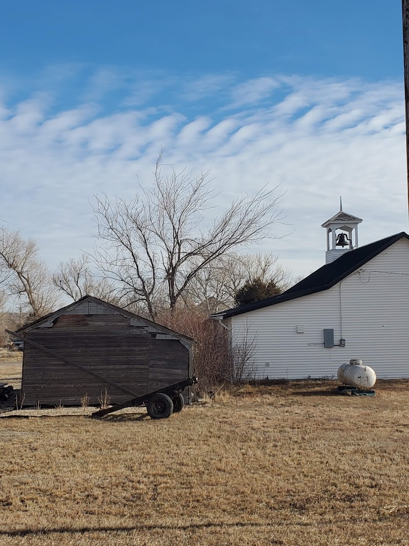 White one-story building with a small bell tower stands beside an old weathered wooden trailer on a dry grassy lot in Badlands National Park.