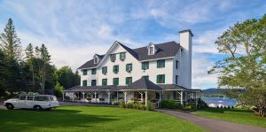 White hotel with green shutters and wraparound porch sits on a manicured lawn beside a lake in Acadia National Park.