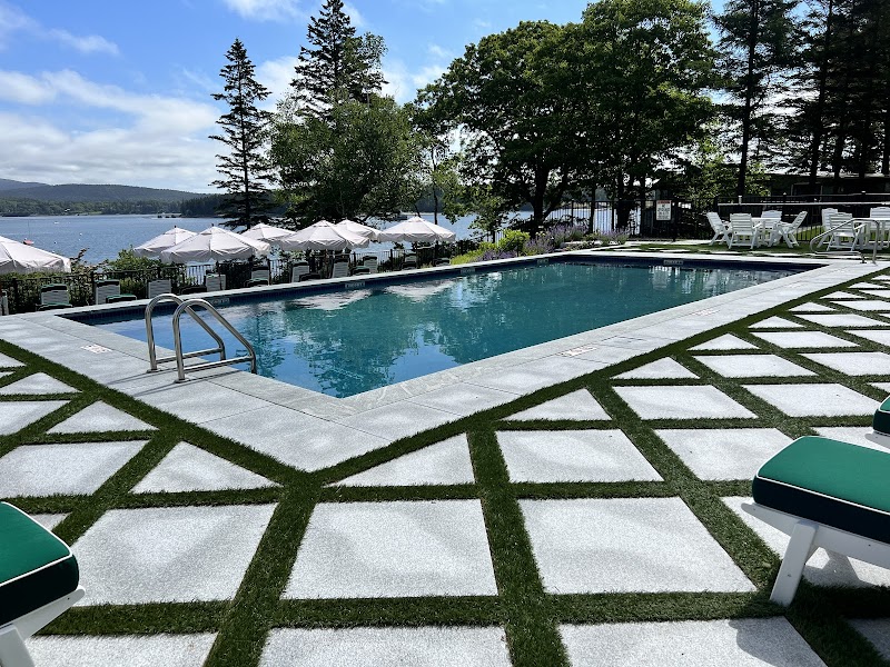 Turquoise pool with white tile surround, geometric grass inlays, and lounge chairs along a lake in Acadia National Park.