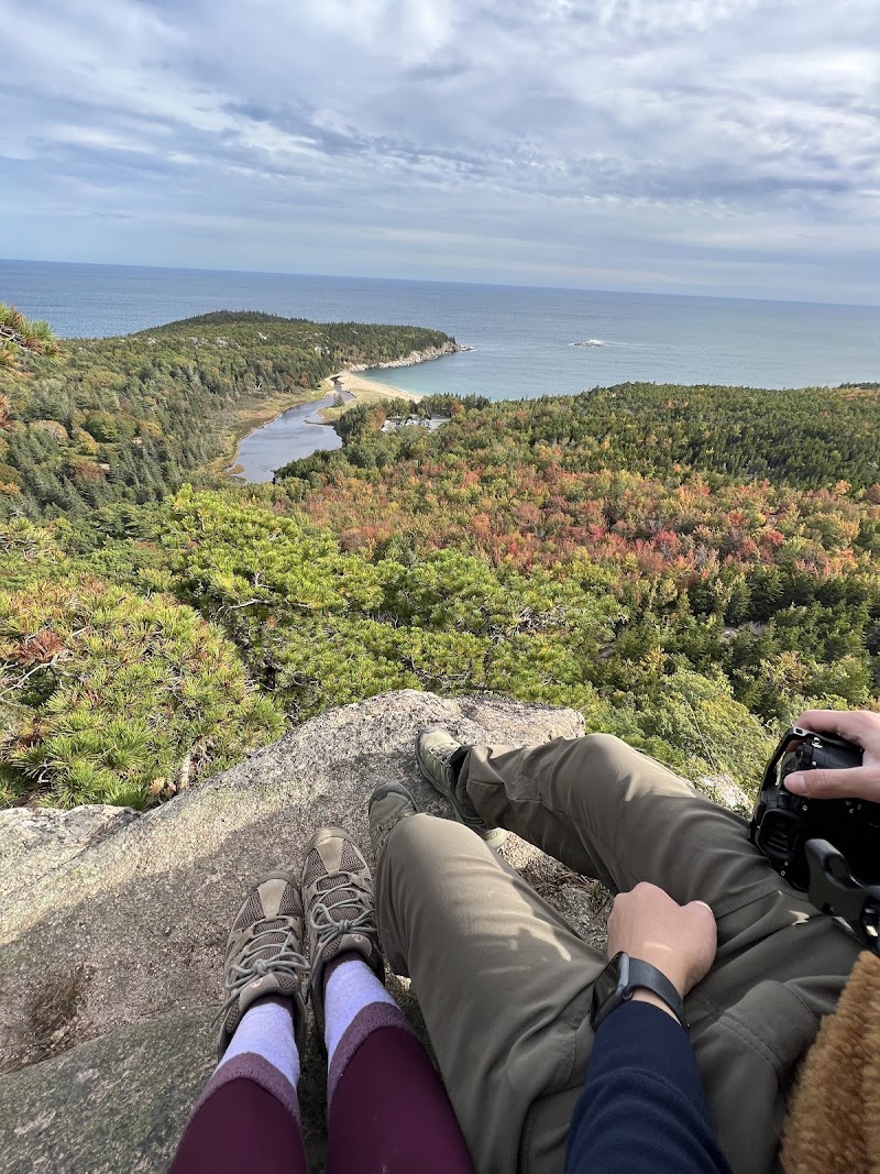 Overlook from Beehive Trail in Acadia National Park; hikers sit on a rock with fall foliage and the blue Atlantic beyond.
