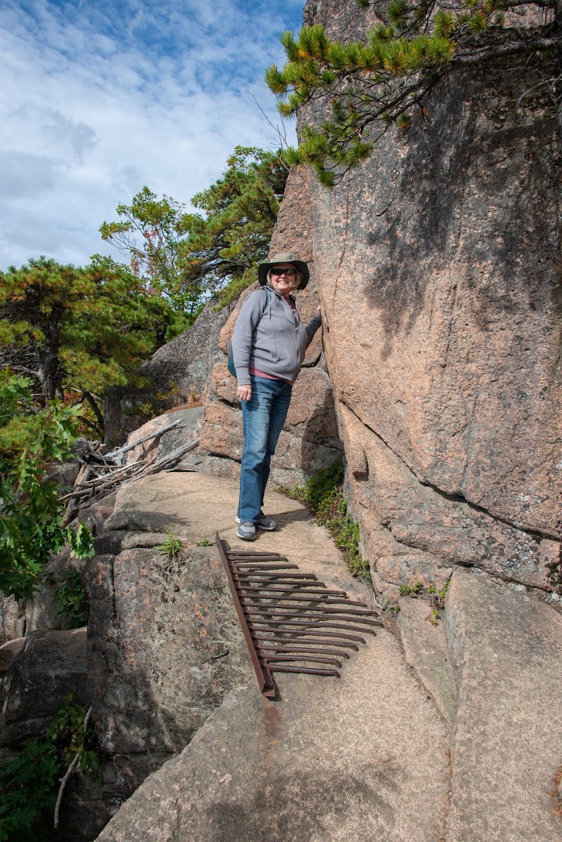 Hiker in a gray jacket and wide-brim hat stands on a rocky ledge beside a granite wall with a metal grate, Acadia National Park.