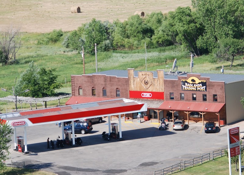 Brick trading post with red canopy and gas pumps, parked cars and motorcycles beside green trees at Badlands National Park.