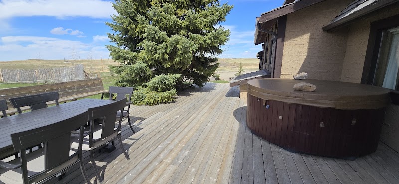 Spacious wooden deck with a black dining table and chairs, a round hot tub by a beige house, and an evergreen tree over Glacier National Park.