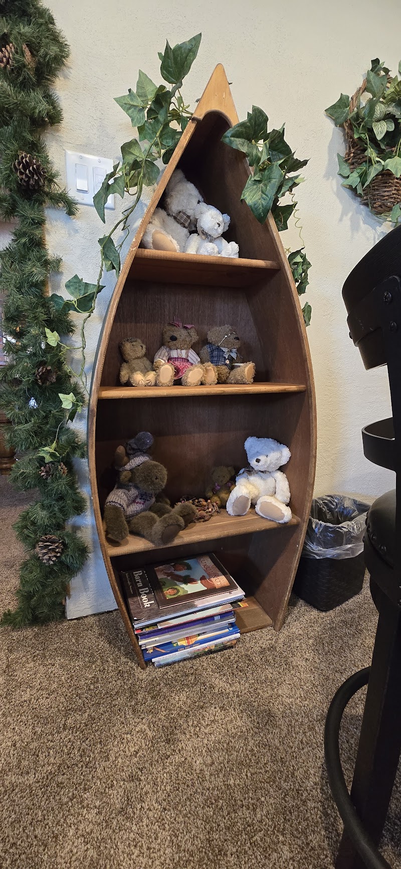 Boat-shaped wooden shelf filled with teddy bears on three shelves, ivy garlands, pinecones, and a stack of books in Glacier National Park.
