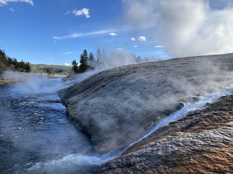 Firehole River winds along steaming geothermal banks in Yellowstone National Park, with blue sky and trees in the background.
