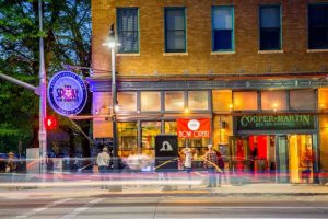 Brick storefronts with glass fronts and neon signs line a busy city street, pedestrians blur by near Arches National Park.