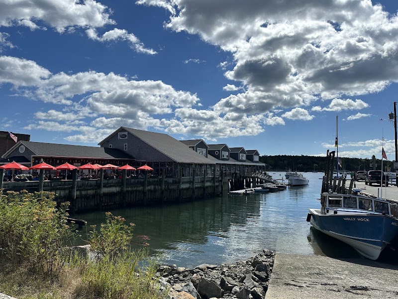 Harbor scene in Acadia National Park showing a pierside building, red umbrellas, and boats docked along calm water.