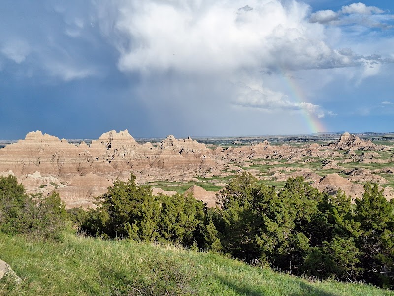 Badlands National Park: layered sandstone spires across a green valley, with trees in foreground and a rainbow in a cloudy sky.