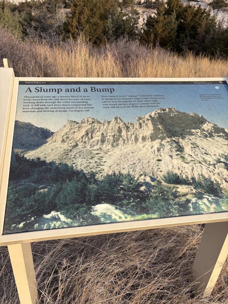 Cliff Shelf Trail sign at Badlands National Park with a photo of jagged limestone hills and dry grasses.
