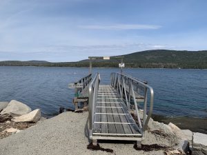 Metal ramp dock extends into a calm blue lake at Acadia National Park, with rocky shore and forested hills beyond.