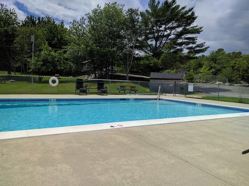 Acadia National Park campground pool area with a bright blue pool, fence, lounge chairs, and picnic tables beside green trees.