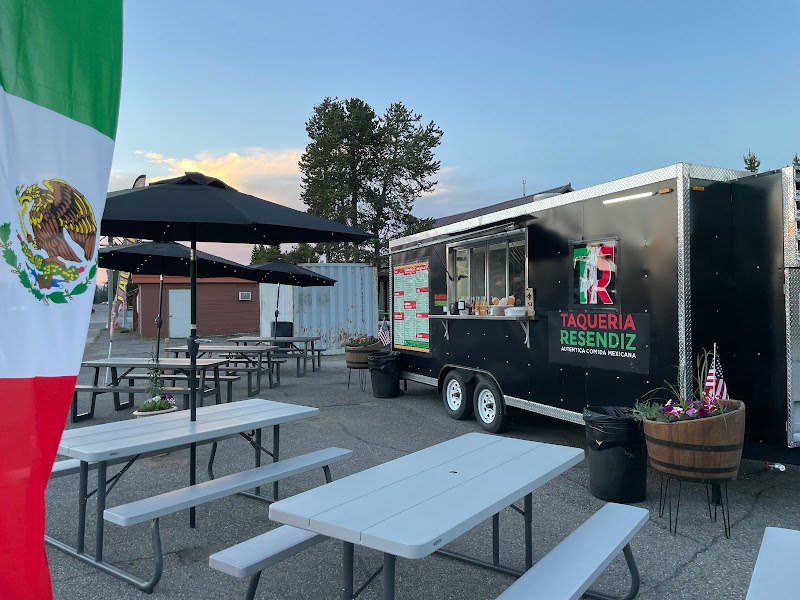 Outdoor seating with gray picnic tables and a black taqueria truck under umbrellas, Mexican flag in Yellowstone National Park.