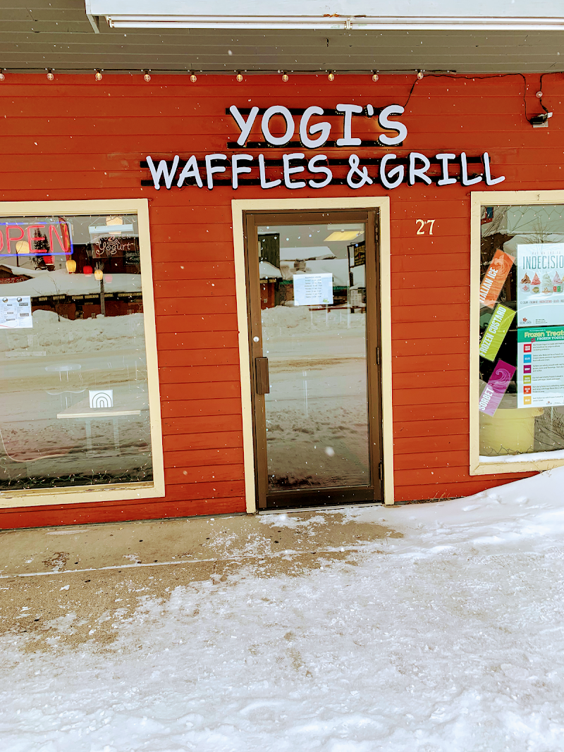 Red wooden restaurant storefront with glass door and large windows in Yellowstone National Park, snow-covered sidewalk and string lights.