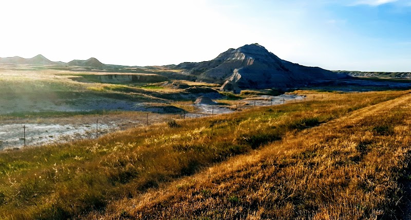 Rolling golden prairie leads to a craggy clay hill, with fences and a shallow reflective wash in Badlands National Park.
