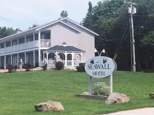 Two-story light gray motel with a white wraparound porch on a grassy lawn near a sign in Acadia National Park.