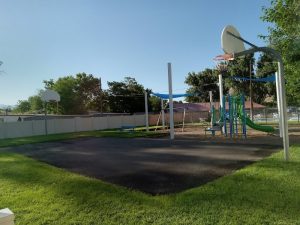 Playground with blue shade sails, swings, a slide, basketball hoop, and a paved court beside green grass at Arches National Park.