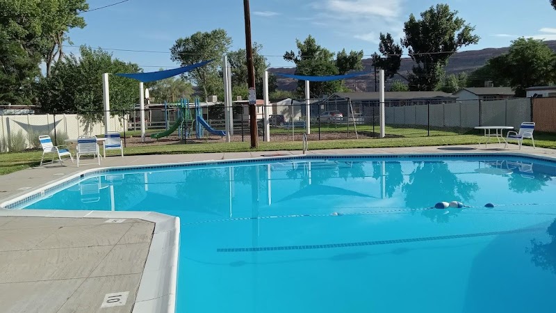 Blue outdoor pool with white lounge chairs and shade sails beside a fenced yard and playground near Arches National Park.