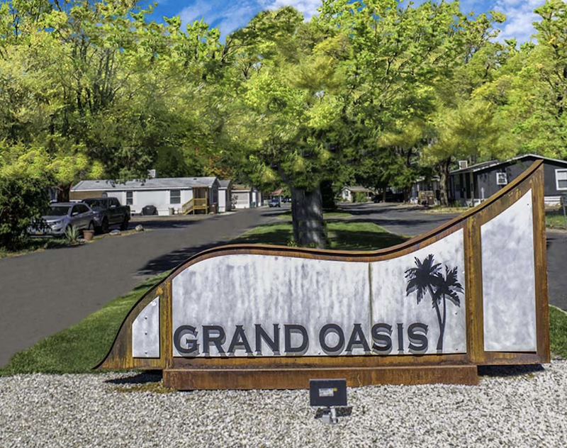 Curved, weathered lodging sign at a campground entrance in Arches National Park, with mobile homes, trees, and a paved road.